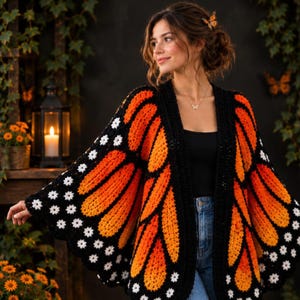 May include: A crochet cardigan featuring a monarch butterfly wing pattern in vibrant orange, black, and white. The garment has wide sleeves, a black border, and small white floral accents along the edges, worn over a black top and blue jeans.
