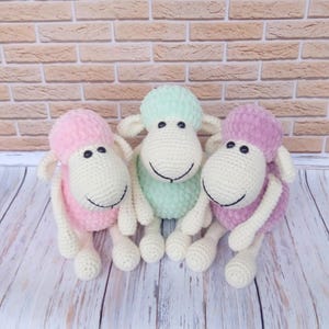 Op de afbeelding: Drie handgemaakte gehaakte schapen in pastelkleuren. Elk schaap heeft een witte kop, lichaam en poten, met een gekleurde kop en lichaam. De schapen zijn naast elkaar geplaatst, met roze, groene en paarse accenten. De achtergrond is een bakstenen muur.