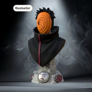May include: A bust of a character with a distinctive orange mask featuring a spiral design. The figure wears a black cloak with a red stripe and has spiky black hair. The base is stone-like with red cloud and symbol details. The word "Bestseller" is in the top left.