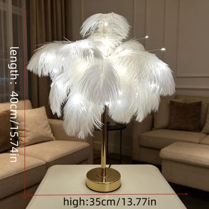 May include: A decorative table lamp featuring a gold base and stem, topped with a cluster of white feathers. The lamp is 15.74 inches (40cm) in length and 13.77 inches (35cm) high. Small lights are interspersed among the feathers, creating a soft glow.