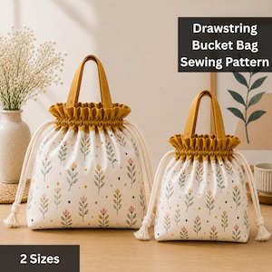 May include: Two drawstring bucket bags in two sizes, featuring a cream-colored fabric with a repeating pattern of small pink and yellow flowers, green leaves, and dark dots. Each bag has a mustard yellow top trim, handles, and white drawstrings.