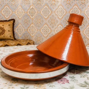 May include: An open, orange-red tagine pot with a conical lid, set on a floral patterned tablecloth. The base of the pot has a white band. The background features a patterned wall and a decorative pillow.