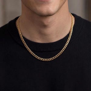 Könnte beinhalten: Eine goldfarbene Panzerkettenhalskette. Die Halskette wird um den Hals getragen und liegt auf einem schwarzen Pullover. Die Kette besteht aus ineinandergreifenden Gliedern und hat eine glänzende Oberfläche. Die Halskette ist ein klassisches Schmuckstück.