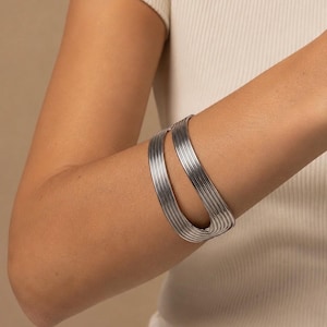 Könnte beinhalten: Ein silberfarbenes Armband in Manschettenform mit einer Reihe paralleler, erhabener Rippen, die ein texturiertes, lineares Muster bilden. Das Armband hat ein offenes Wickeldesign, das eine breite, geschwungene Form um das Handgelenk bildet.