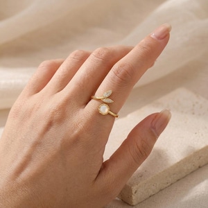 Könnte beinhalten: Ein goldfarbener Ring mit Blattdesign und einem kleinen, runden, weißen Stein. Der Ring hat ein zartes, offenes Design und wird am Finger getragen. Der Ring ist ein Schmuckstück.