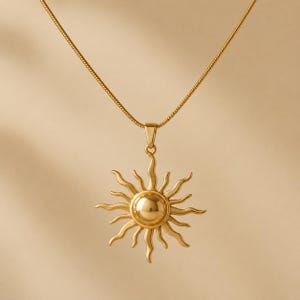 Könnte beinhalten: Eine goldfarbene Halskette mit einem Sonnenanhänger. Der Anhänger hat eine zentrale Kugel mit strahlenförmigen Strahlen. Die Kette ist ein filigranes, dünnes Design. Die Halskette wird vor einem neutralen Hintergrund präsentiert.