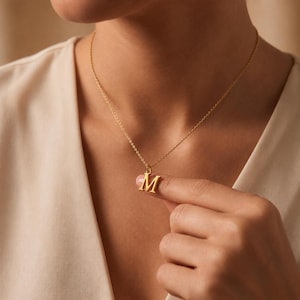 Könnte beinhalten: Eine goldfarbene Halskette mit einer feinen Kette und einem einzelnen Anhänger in Form des Großbuchstabens M. Der Anhänger ist an einem kleinen Verbindungsring an der Kette befestigt und weist durchgehend eine polierte Metalloberfläche auf.