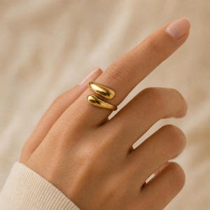 Könnte beinhalten: Ein goldfarbener offener Ring mit zwei glatten, abgerundeten, tropfenförmigen Enden, die sich zueinander biegen. Das Metall hat eine polierte, reflektierende Oberfläche, die für ein elegantes und durchgehendes Erscheinungsbild am Finger sorgt.