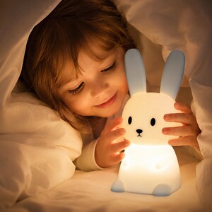 Puede incluir: Una suave y brillante luz de noche con forma de conejito blanco con orejas y pies azules. La luz la sostiene un niño bajo una manta blanca. El conejito tiene ojos negros y una pequeña nariz negra.