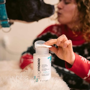 May include: A white cylindrical bottle of Dogmoxie Anytime Anywhere Tonic for paws with a clear cap and a white cotton pad resting on top. The label features black text and blue paint splatter graphics, indicating cleansing and skin barrier care.