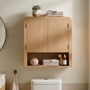 May include: A light oak wood wall-mounted bathroom cabinet featuring two doors with rounded top corners and a lower open shelf. The cabinet is mounted above a toilet and contains folded towels, a small succulent, and a glass bottle.