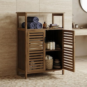 May include: A brown wooden bathroom cabinet with an open top shelf and two slatted doors. The top shelf holds three rolled blue towels, a glass reed diffuser, and a small tray. The interior shelves contain white bottles, a woven basket, and stones.