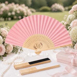 May include: A pink folding hand fan with a light wood frame featuring laser-engraved text T & L, Tania & Liam, and the date 11.12.2026. Two additional folded fans, one with black fabric and one with white fabric, lie in front of the open fan.