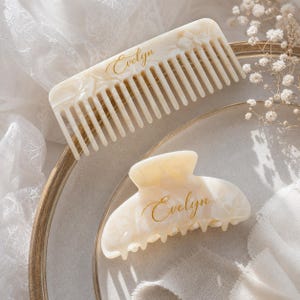 May include: A cream-colored hair comb and a matching hair claw clip, both featuring a marbled pattern and the name Evelyn printed in gold cursive script. The hair accessories are made of a smooth, polished material with a glossy finish.