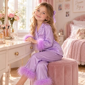 Könnte beinhalten: Ein helllila Satin-Pyjama-Set bestehend aus einem langärmeligen Oberteil mit Knopfleiste und einer passenden Hose. Sowohl die Ärmelbündchen als auch die Hosenbeinsäume sind mit passenden lila Federn besetzt. Das Oberteil zeigt den Namen August mit Herz.