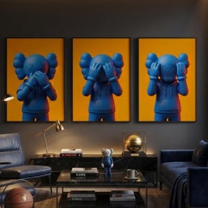 May include: Three framed wall art prints featuring blue KAWS-style figures against a solid yellow background. The figures are depicted in the see-no-evil, hear-no-evil, and speak-no-evil poses, each wearing a hooded sweatshirt with X-shaped eyes.