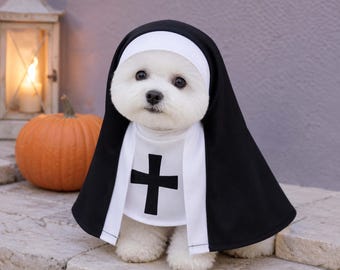 May include: A small white dog wearing a nun costume consisting of a black habit with a white headpiece and a white chest panel featuring a black cross symbol. The costume covers the head and torso, creating a traditional religious appearance.