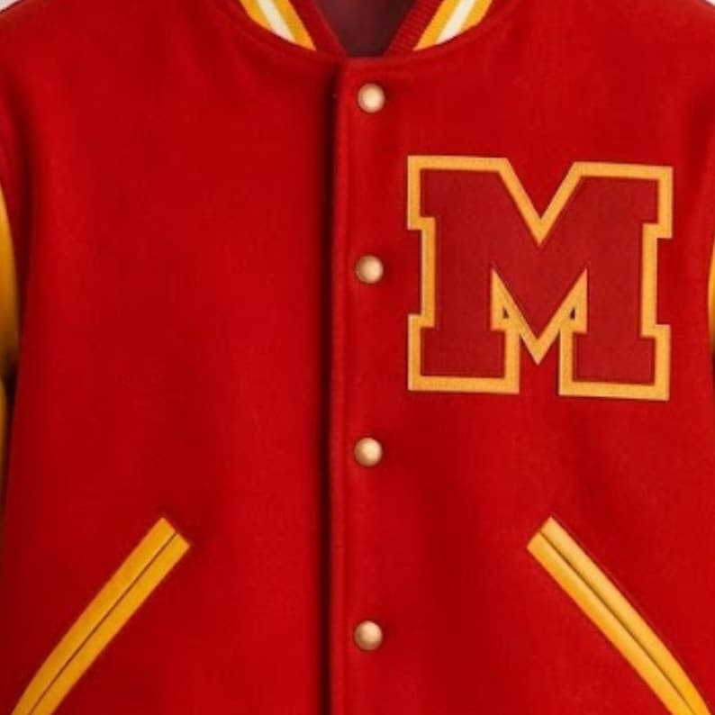 Puede incluir: Una chaqueta universitaria roja con un gran parche de la letra M en rojo oscuro con borde amarillo en el pecho. La chaqueta tiene cierre frontal de botones met&aacute;licos, cuello con rayas amarillas y detalles amarillos en los bordes de los bolsillos.