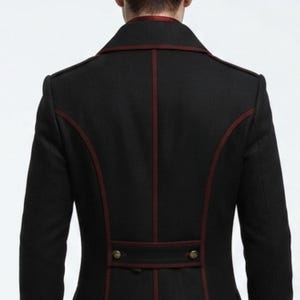 May include: A black jacket featuring dark red piping along the seams, a high collar, and a horizontal back tab detail with two metallic buttons. The garment is shown from the back, highlighting the structured paneling and the deep red trim.