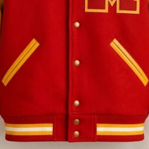 Puede incluir: Una chaqueta universitaria roja con una letra M amarilla en el pecho. La chaqueta tiene un cierre frontal con botones a presi&oacute;n dorados, dos bolsillos diagonales con ribete amarillo y un dobladillo acanalado con rayas horizontales amarillas y blancas.