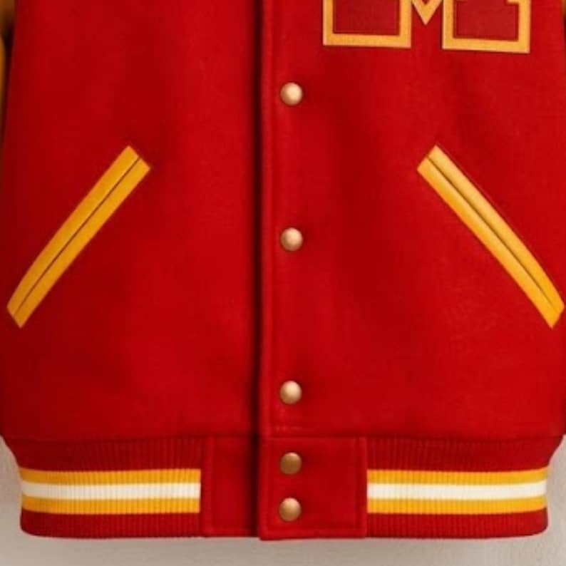Puede incluir: Una chaqueta universitaria roja con una letra M amarilla en el pecho. La chaqueta tiene un cierre frontal con botones a presi&oacute;n dorados, dos bolsillos diagonales con ribete amarillo y un dobladillo acanalado con rayas horizontales amarillas y blancas.