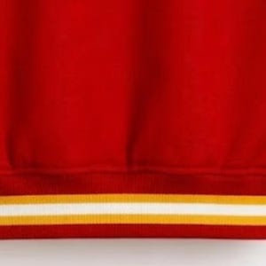 Puede incluir: Una sudadera de color rojo brillante con un dobladillo acanalado en la parte inferior. El dobladillo presenta dos rayas horizontales, una de color amarillo dorado y otra blanca, que corren paralelas a lo largo del borde de la tela.