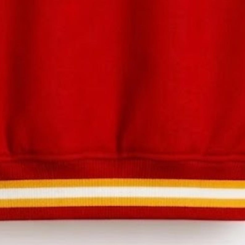 Puede incluir: Una sudadera de color rojo brillante con un dobladillo acanalado en la parte inferior. El dobladillo presenta dos rayas horizontales, una de color amarillo dorado y otra blanca, que corren paralelas a lo largo del borde de la tela.