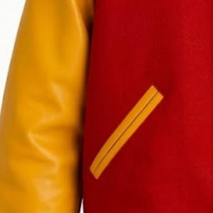 Puede incluir: Una chaqueta universitaria con un cuerpo de lana de color rojo vibrante y mangas de color amarillo mostaza en contraste. La prenda incluye un bolsillo de ojal inclinado en la parte delantera, ribeteado con material amarillo mostaza a juego.
