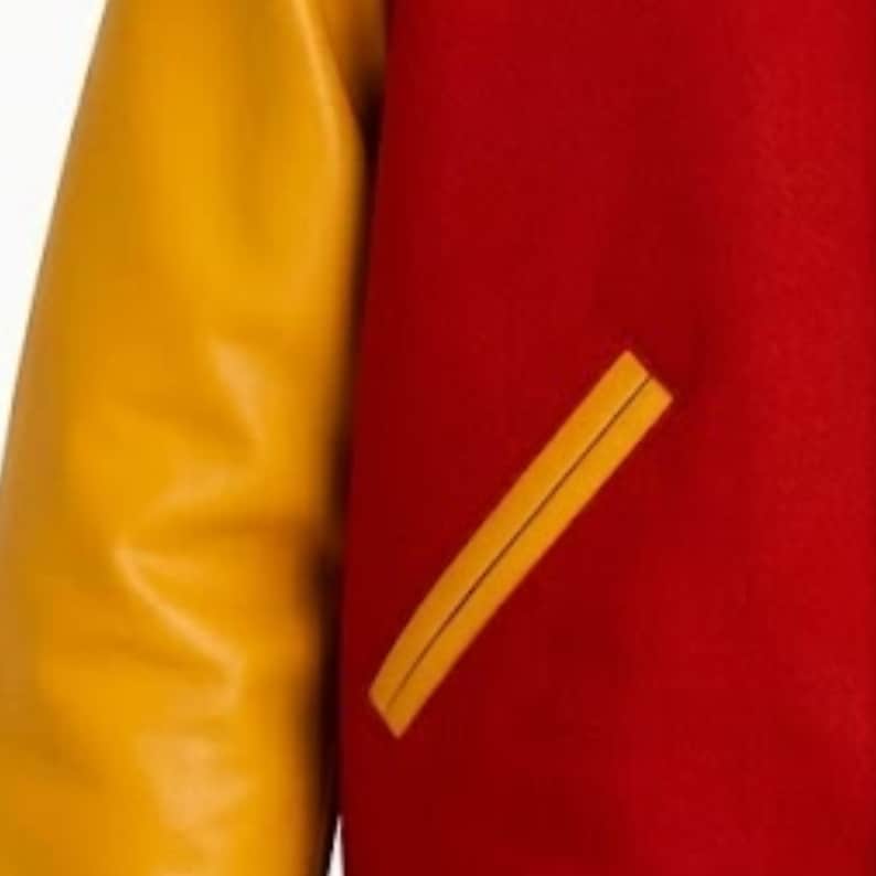 Puede incluir: Una chaqueta universitaria con un cuerpo de lana de color rojo vibrante y mangas de color amarillo mostaza en contraste. La prenda incluye un bolsillo de ojal inclinado en la parte delantera, ribeteado con material amarillo mostaza a juego.