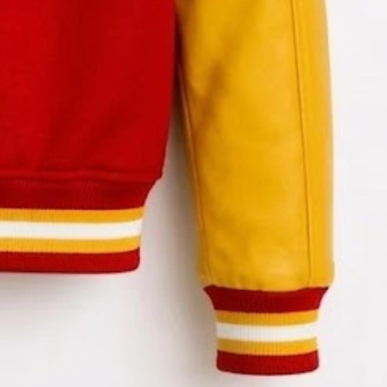 Puede incluir: Una chaqueta universitaria con cuerpo rojo y mangas amarillas en contraste. Los pu&ntilde;os y el dobladillo presentan un borde de punto acanalado con rayas horizontales en rojo, blanco y amarillo, ofreciendo una est&eacute;tica deportiva.
