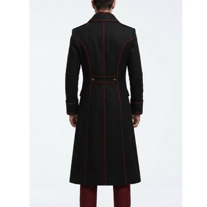 May include: A long black coat featuring structured seams highlighted by thin dark red piping. The coat includes a high collar, long sleeves with cuffs, and a horizontal back tab detail with two small metallic buttons at the waistline.
