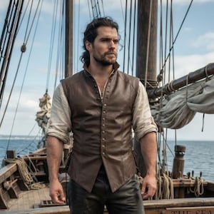 May include: A person with dark hair and a beard wearing a brown leather vest over a light-colored long-sleeved shirt with rolled-up cuffs and dark trousers, standing on the wooden deck of a sailing ship with ropes and masts visible in the background.