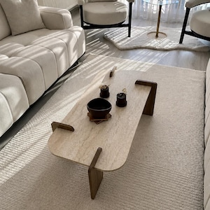 Peut inclure: Une table basse avec un plateau rectangulaire en pierre travertin aux coins arrondis et des pieds en bois brun foncé qui s'insèrent dans la surface. Sur la table se trouvent un bol brun foncé et deux petits récipients cylindriques.