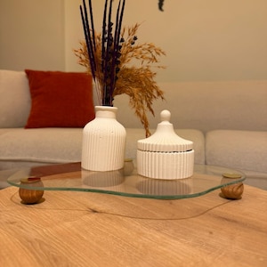 Könnte beinhalten: Ein dekoratives Glastablett mit Holzfüßen trägt eine weiße Keramikvase und ein Gefäß mit Deckel. Die Vase enthält getrocknete lila und beige Blumenarrangements. Das Tablett steht auf einem hellen Holztisch.