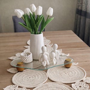 Puede incluir: Un jarrón blanco con tulipanes blancos sobre una mesa de madera. La mesa está decorada con una bandeja de cristal, salvamanteles de ganchillo y figuras blancas. También hay un portavelas blanco con un acento dorado.