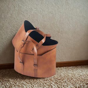 Könnte beinhalten: Zwei gestapelte, braune Leder-Tragetaschen mit schwarzem Innenraum. Die Taschen haben Griffe und verstellbare Riemen. Die Taschen stehen auf einem beigefarbenen Teppich, mit einer beigefarbenen Wand im Hintergrund.