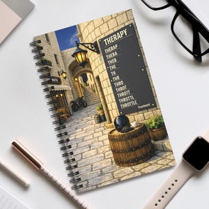 May include: Spiral-bound notebook featuring a detailed illustration of a European street scene with a motorcycle. The cover includes the word "THERAPY" and a list of words. A pen, watch, and glasses are also visible.