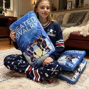 May include: A rectangular blue pillow featuring the text HEATED RIVALRY and an illustration of two hockey players facing each other on ice. The pillow is held by a person wearing a navy blue long-sleeve shirt with a hockey graphic and matching pants.
