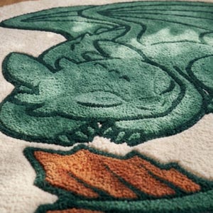 Può includere: Un tappeto rotondo con un design di drago verde e dettagli arancioni. Il drago è raffigurato in stile cartone animato, con ali dettagliate e un'espressione amichevole. Il tappeto ha uno sfondo color crema.