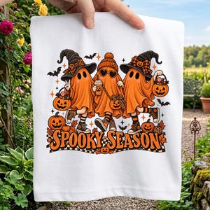 Może przedstawiać: Biały t-shirt z grafiką trzech pomarańczowych duchów w kapeluszach i dodatkach, trzymających wiaderka w kształcie dyń oraz napój. Wzór zawiera nietoperze, gwiazdki i napis "SPOOKY SEASON" w kolorze pomarańczowym na czarno-białej szachownicy.