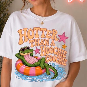 Puede incluir: Camiseta blanca con el texto "Hotter Than a Hoochie Coochie" en naranja y rosa. El gráfico presenta un caimán verde con gafas de sol en forma de corazón, relajándose en un flotador inflable rosa y naranja en el agua.