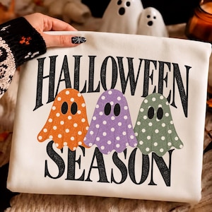 Könnte beinhalten: Ein weißes Sweatshirt mit dem schwarzen Schriftzug HALLOWEEN SEASON und drei Geistern in Orange, Lila und Grün, die jeweils mit einem weißen Punktmuster verziert sind. Die Geister befinden sich zwischen den beiden Textzeilen auf der Vorderseite.