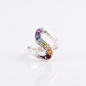 May include: A silver ring shaped like the letter S, featuring a channel set with a row of small, round, multi-colored gemstones including purple, blue, light blue, green, yellow, orange, and red stones arranged in a vibrant gradient pattern.