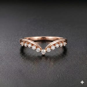 May include: Rose gold ring with a curved design, set with multiple small, round, clear gemstones. The ring has a V-shape, with the gemstones arranged along the curve. The ring is on a dark background.