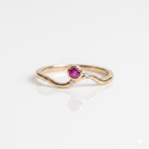 May include: A gold ring featuring a wavy band design with a single round bezel-set pink ruby as the central focal point, accented by two small clear round-cut diamonds set into the band on either side of the ruby in a delicate arrangement.