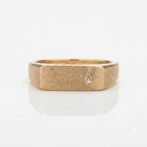 May include: A gold signet ring with a brushed matte finish featuring a single small kite-shaped diamond set off-center on the flat rectangular face. The band has a solid construction with a consistent width and a warm yellow gold tone.