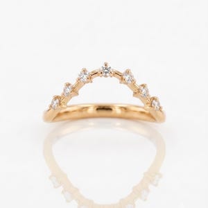 May include: A gold ring featuring a curved, chevron-shaped band set with seven small, round, clear stones. The stones are arranged in a symmetrical arc along the top portion of the polished gold band, creating a pointed, crown-like silhouette.