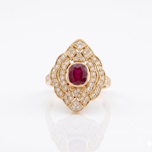 May include: A gold ring featuring a central cushion-cut deep red ruby surrounded by a decorative frame of small round white diamonds. The metal setting has a textured, intricate design that extends outward in a pointed, elongated shape.