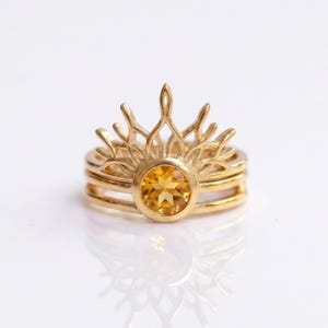 Op de afbeelding: Een gouden ring met een centrale ronde gele citrien edelsteen in een zetting. De band is aan de bovenzijde voorzien van een ingewikkeld, opengewerkt zonnestraalontwerp dat vanaf de steen omhoog straalt en een kroonachtig silhouet vormt.