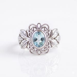Puede incluir: Un anillo de tono plateado con una piedra preciosa de color azul claro de corte ovalado en el centro. El engaste presenta intrincados adornos metálicos en forma de volutas y motivos de hojas detallados que se extienden hacia los lados.